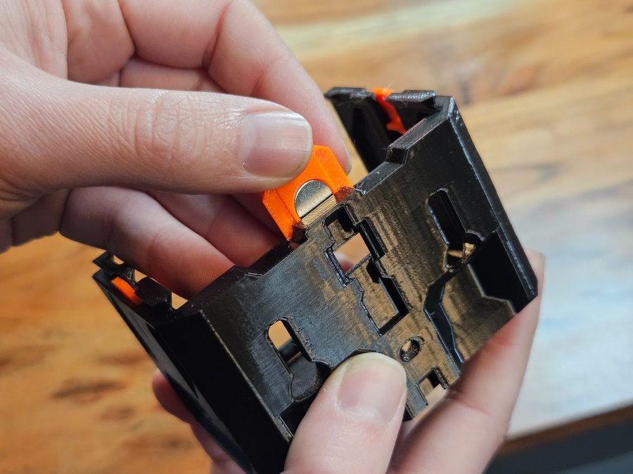 A thumb pressing an orange magnet clip, a round magnet seated in it, into its slot in a small black 59 1W-1H case