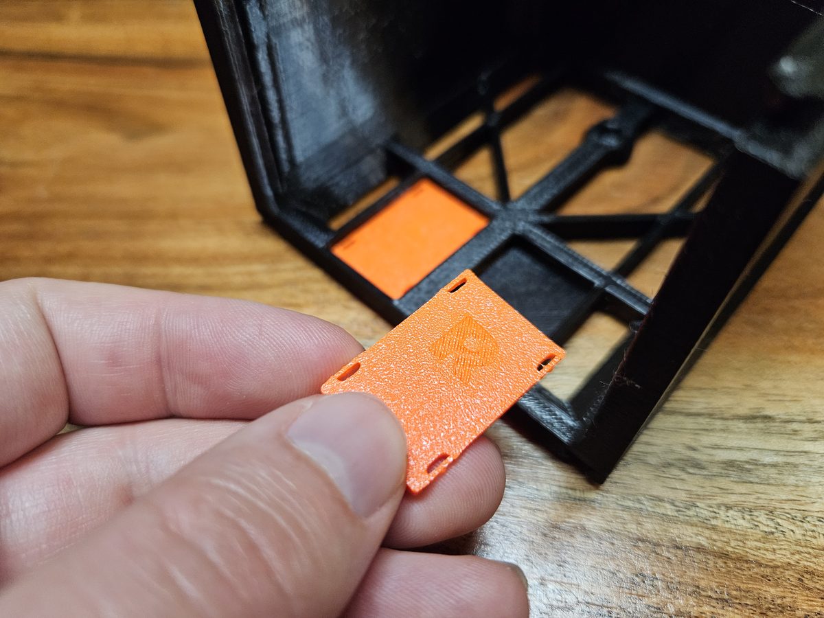 Fingers holding a small flat orange drawer stopper marked R; a second stopper is already snapped into a slot in the floor of the black case behind it