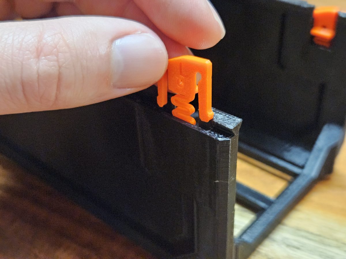 A thumb and finger lowering an orange QuickLock, its coiled spring end down, into the slot in the top of a black case wall; a second QuickLock is already seated in the far wall