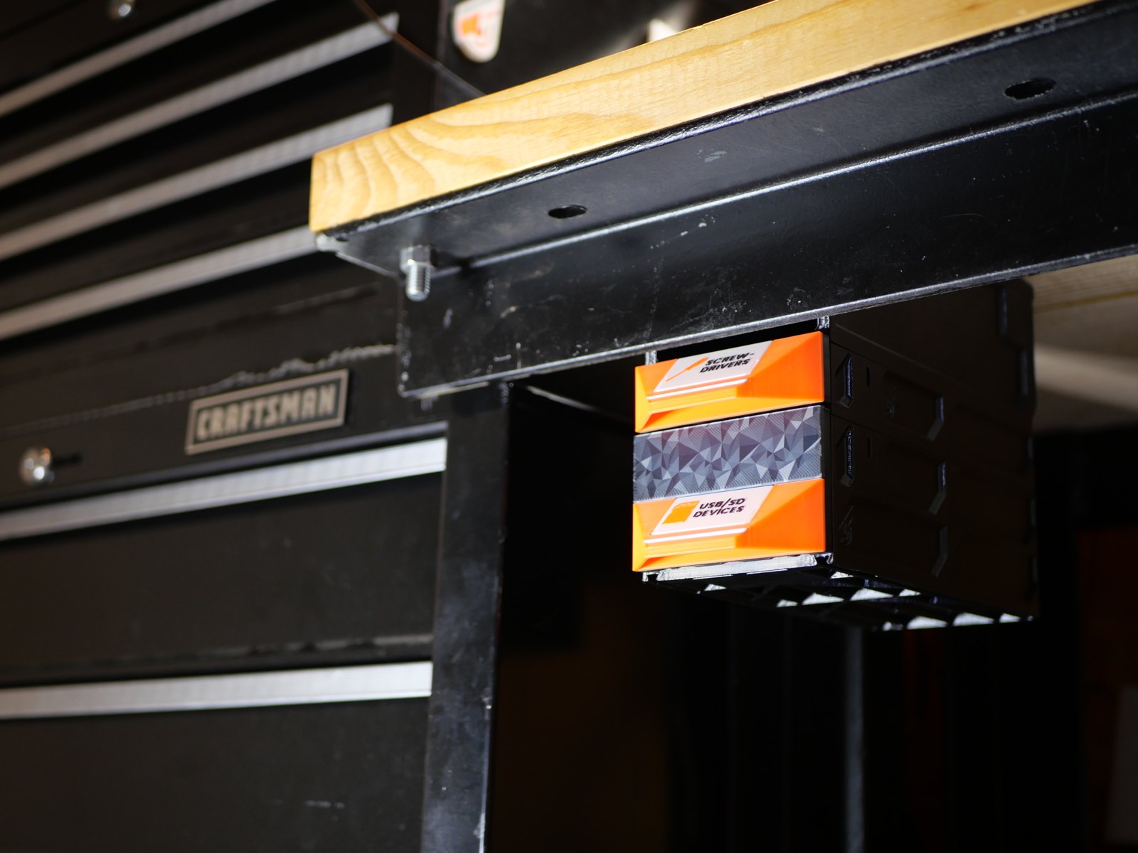 Two stacked cases with orange and gray drawer fronts hanging from an under-table rail beneath the steel lip of a wooden-topped workbench