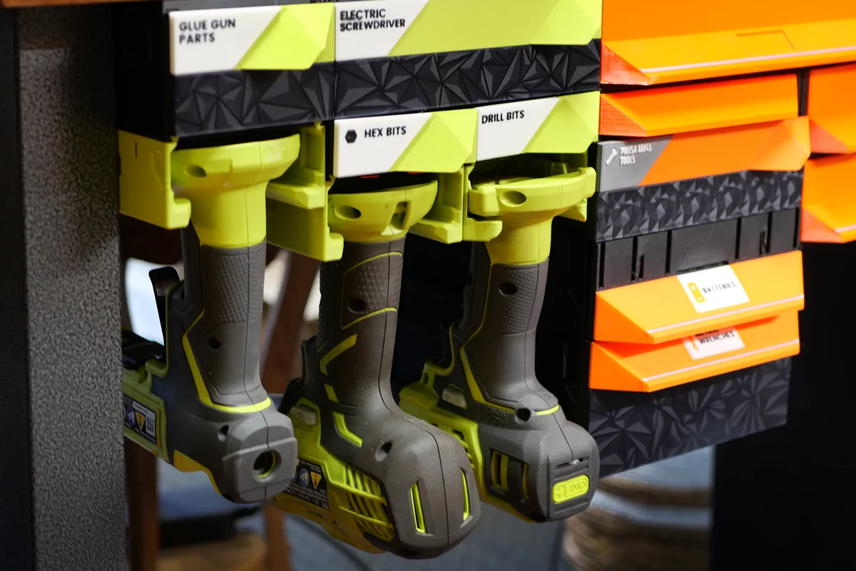 Three cordless power tools hanging in printed holders under a wooden workbench, beneath labeled MODULITH drawers for hex bits, drill bits and glue gun parts, with orange drawers beside them.